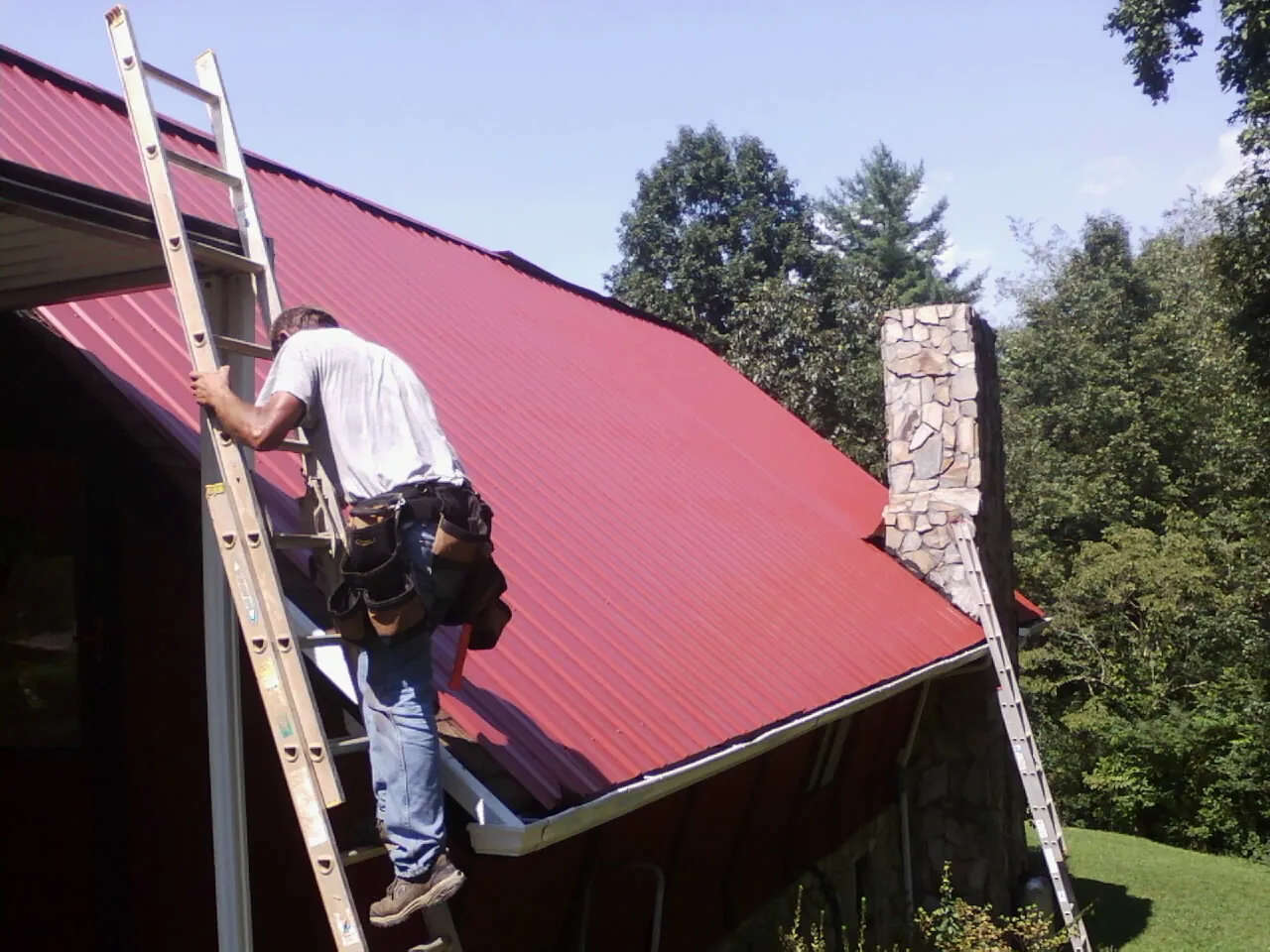 Roofer on a ladder beside a red metal roof and stone chimney — Soffit & Fascia Repair by Roofing Brothers in Harrisonburg